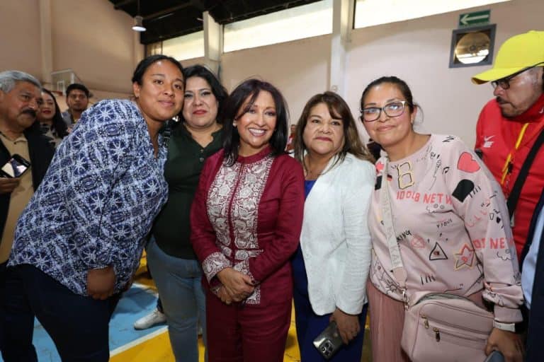 Mujer sonriente en grupo de personas en un evento público, con fondo de interior y varias personas observando.