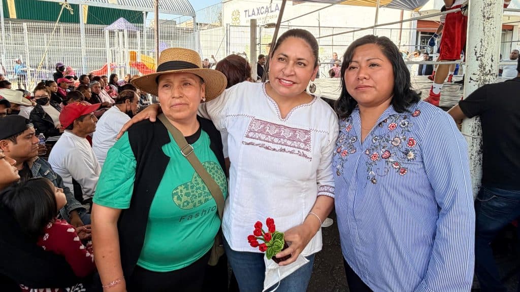 Tres mujeres en un evento público en Tlaxcala, una de ellas sostiene un ramo de flores rojas, rodeadas de muchas personas en un ambiente festivo y comunitario.