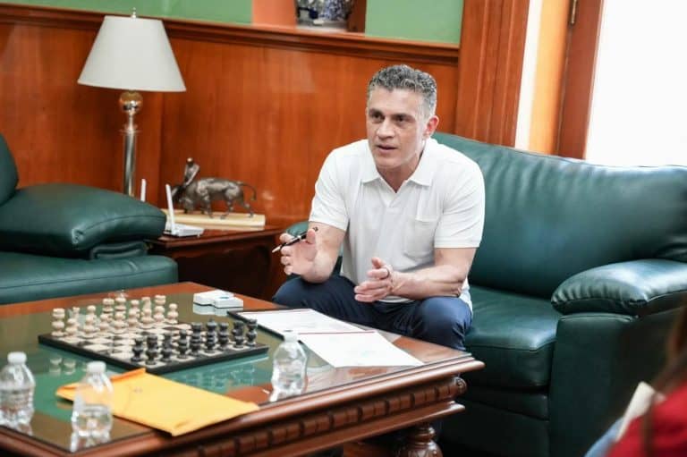 Hombre en una sala de reuniones jugando ajedrez, sentado en un sillón verde, con fondo de madera y una lámpara de pie, en un entorno formal y profesional.