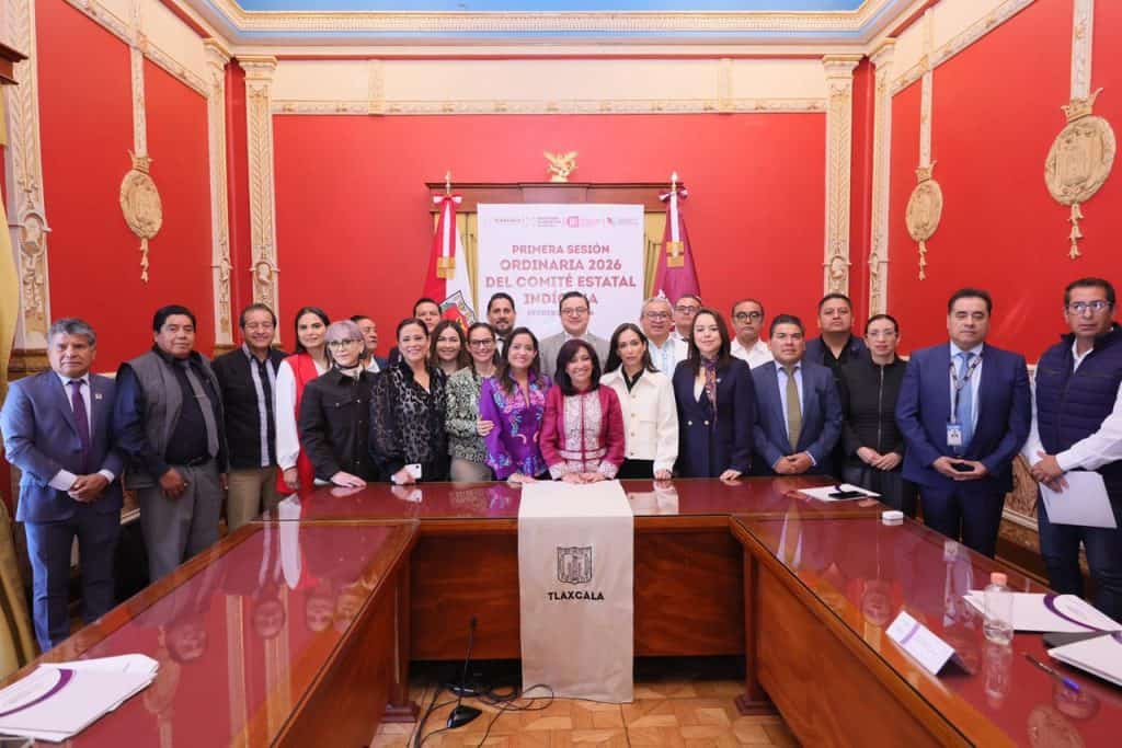 Grupo de funcionarios y representantes en una reunión oficial en un salón con decoración roja y detalles dorados, en Tlaxcala, México.