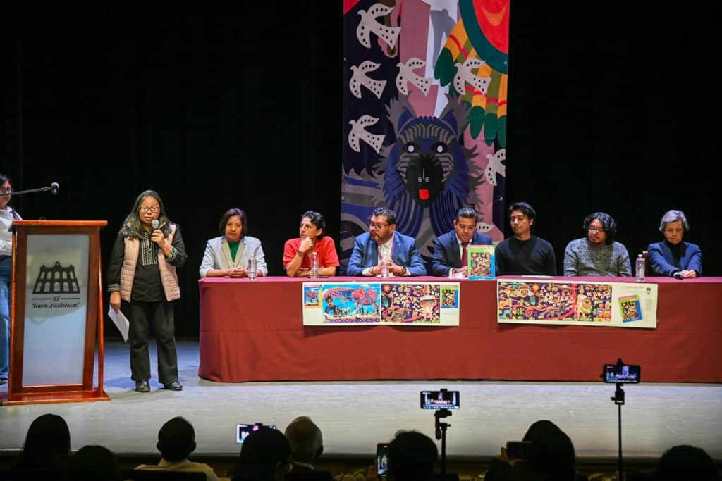 Panel de discusión en un evento cultural con varias personas sentadas en una mesa y una mujer hablando en un micrófono en un escenario con arte colorido de fondo.