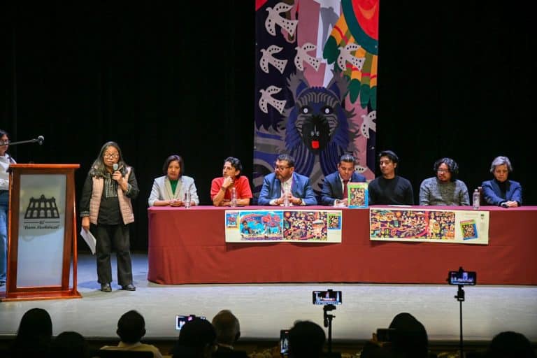 Panel de discusión en un evento cultural con varias personas sentadas en una mesa y una mujer hablando en un micrófono en un escenario con arte colorido de fondo.