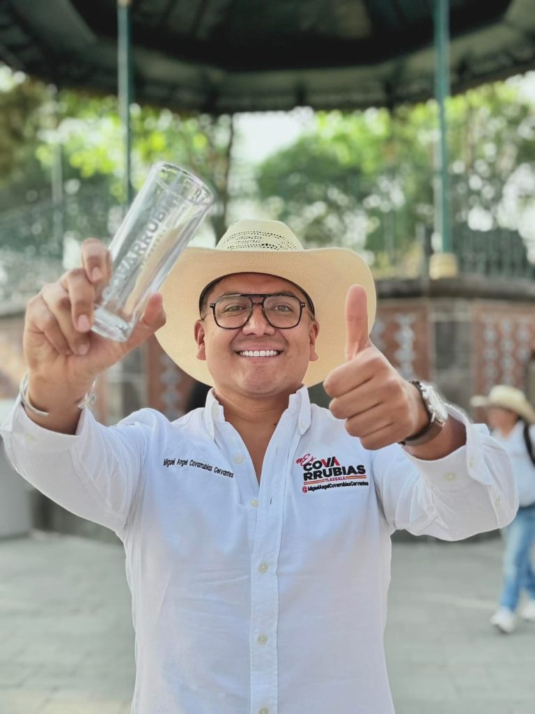 Borracho sonriente con sombrero y gafas, sosteniendo un vaso de cristal y haciendo un gesto de pulgar hacia arriba en un evento al aire libre en Tlaxcala, México.