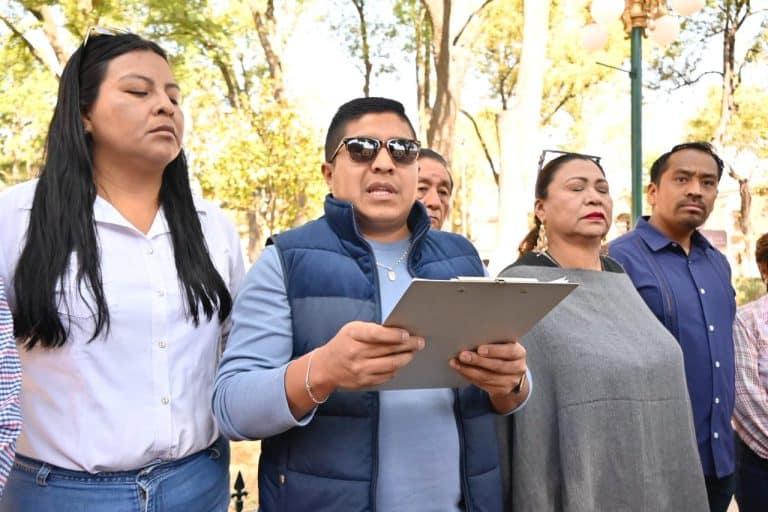 Persona leyendo un documento en un evento público al aire libre, con árboles y farolas en el fondo, en un día soleado y con clima agradable.