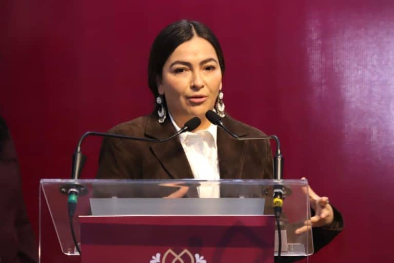 Mujer hablando en un podio durante una conferencia o evento formal, con fondo rosa y micrófonos frente a ella.