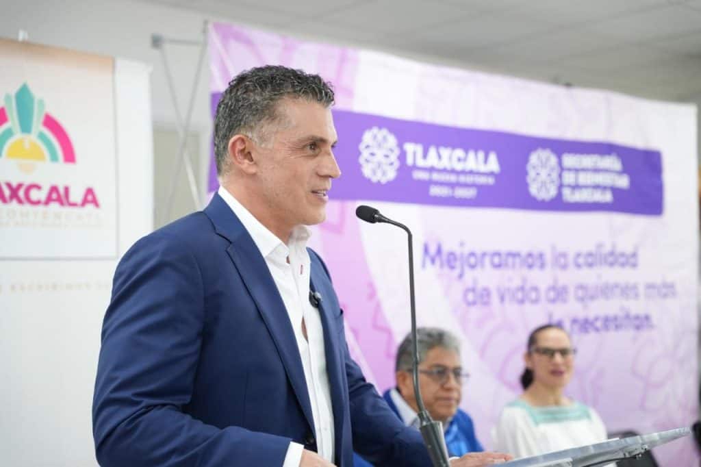 Hombre en traje azul hablando en un evento oficial con fondo de banner morado y personas en la mesa de discusión.