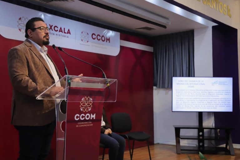 El hombre en un evento de comunicación en un escenario con pantalla y logotipos de Tlaxcala y CCOM.