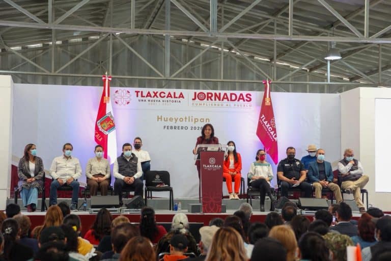 La mujer en el podio durante la conferencia en un evento en Tlaxcala, con varias personas sentadas en el escenario y una audiencia en primer plano.