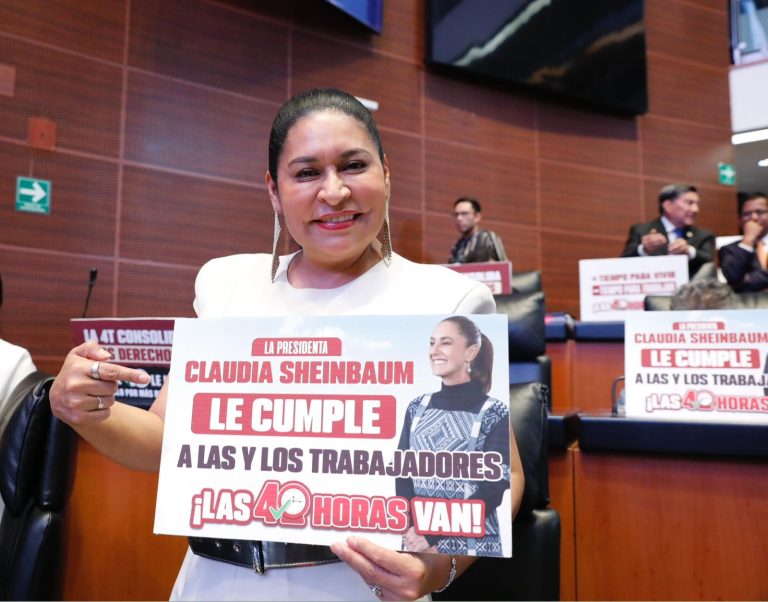 Mujer sosteniendo cartel en evento político, celebrando el cumplimiento de las 40 horas laborales, en un entorno formal con otros asistentes en el fondo.
