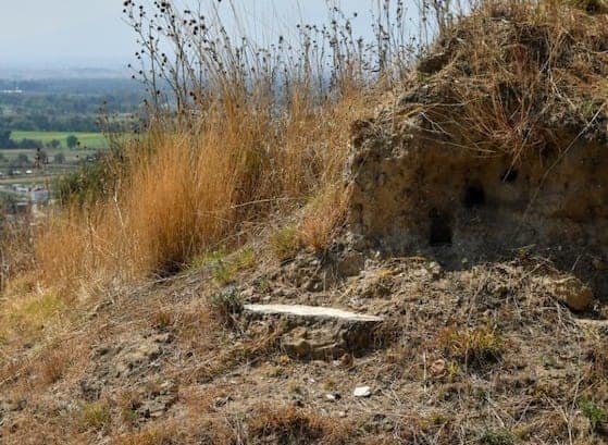 Restos de una estructura antigua en un terreno árido con vegetación seca y vista panorámica en el fondo.
