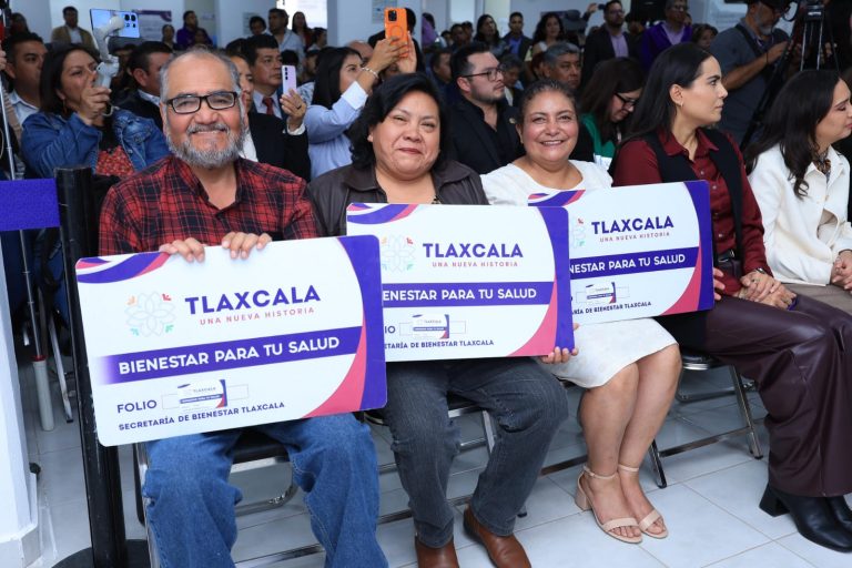 Personas sonriendo y sosteniendo carteles con el logo del gobierno de Tlaxcala en un evento de bienestar para la salud. La audiencia participa en una conferencia o ceremonia oficial en un espacio cerr.