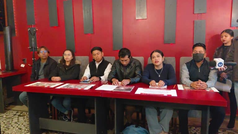 Audiencia de prensa con varios periodistas y representantes en una mesa, en un evento formal en un espacio con paredes rojas y decoraciones modernas.