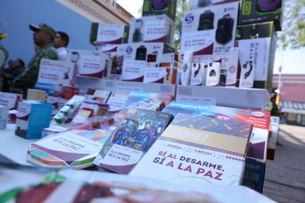 Montón de productos y folletos en una mesa, incluyendo libros y artículos promocionales, en un evento público en Tlaxcala, México.