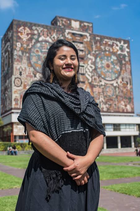 Mujer sonriendo frente a un mural colorido en un parque, vestida con ropa oscura y bufanda, en Tlaxcala, México.