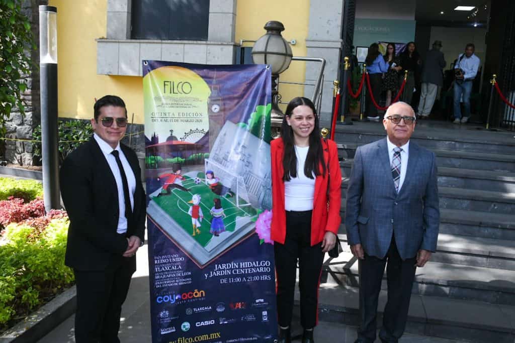 Tres personas en la entrada de un edificio, con un cartel promocional de un evento cultural en Tlaxcala, México.
