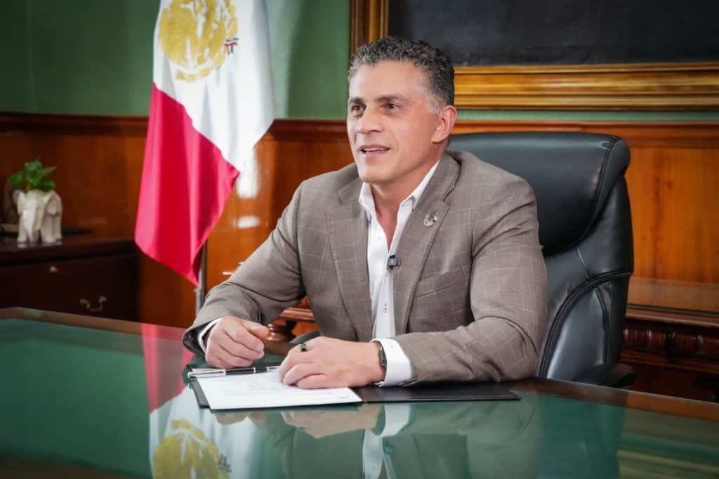 Hombre en una oficina con bandera de México al fondo, sentado en una mesa de vidrio, vestido con traje gris y camisa blanca, en un entorno formal y profesional.