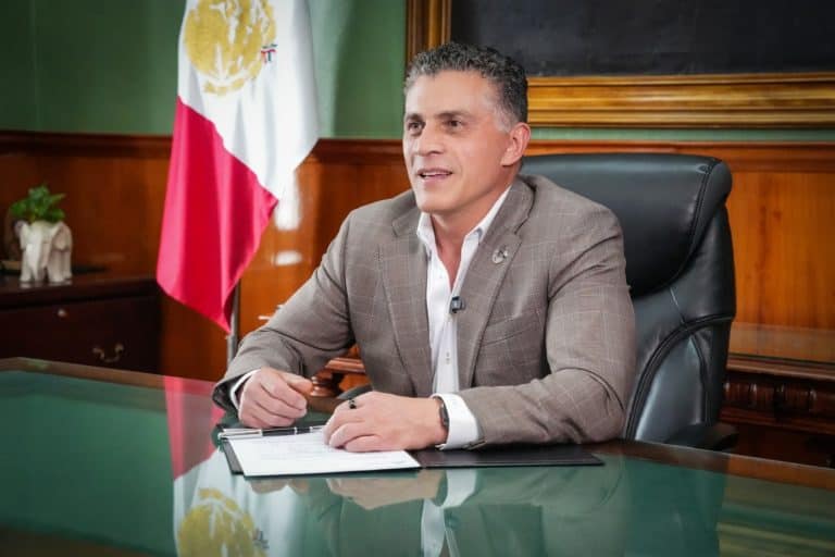 Hombre en una oficina con bandera de México al fondo, sentado en una mesa de vidrio, vestido con traje gris y camisa blanca, en un entorno formal y profesional.
