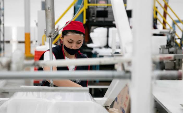 Cinta de producción en una fábrica con una trabajadora operando maquinaria en un entorno industrial.