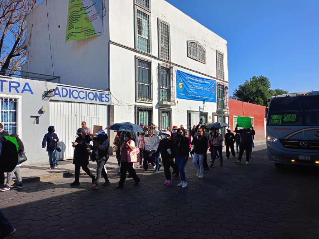 Personas haciendo fila afuera de un edificio en un día soleado, algunos con paraguas, en una calle con autobús y árboles al fondo.