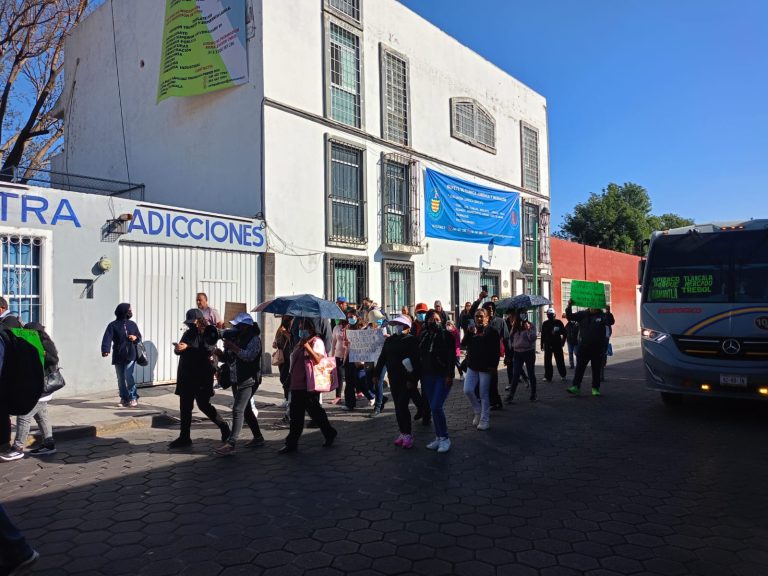 Personas haciendo fila afuera de un edificio en un día soleado, algunos con paraguas, en una calle con autobús y árboles al fondo.