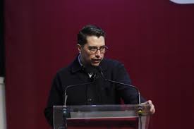 El hombre con gafas y barba, vestido con chaqueta negra, hablando en un podio con fondo rojo.
