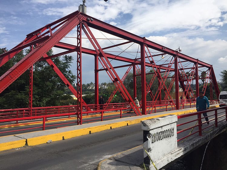 Puente peatonal y vehicular de estructura metálica roja sobre un río, con cielo nublado y personas caminando en la acera.
