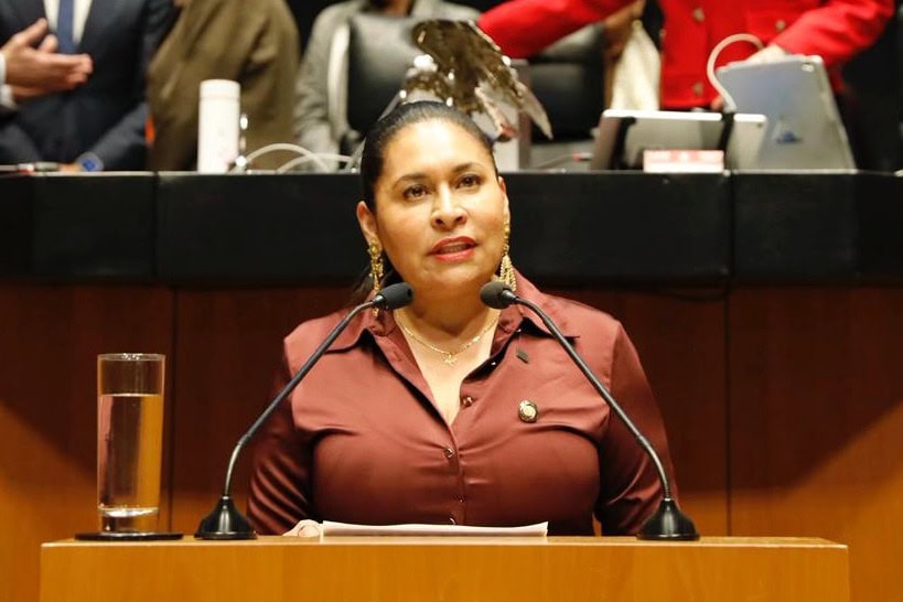 Mujer en un podio durante una conferencia o sesión en un entorno formal, con micrófonos y asistentes al fondo.