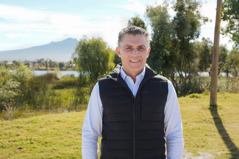 Hombre sonriendo al aire libre en un parque con árboles y montañas al fondo, vestido con camisa blanca y chaleco negro, en un día soleado y despejado.