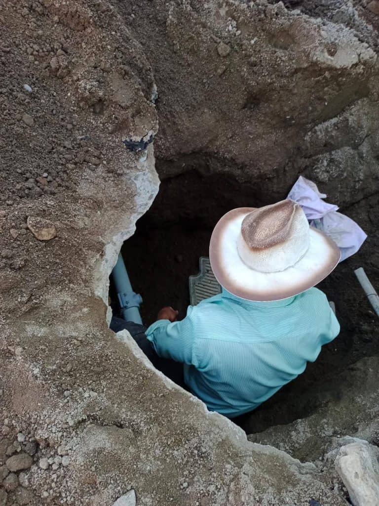 Persona con sombrero sentado en un hoyo en la tierra, posiblemente realizando trabajo de excavación o investigación en un entorno al aire libre.