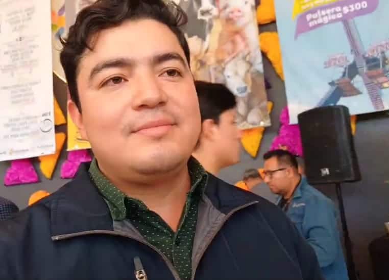 Persona joven con cabello oscuro y piel clara, vistiendo una chaqueta azul y camisa de color verde con puntos blancos, en un evento público con decoraciones coloridas y otras personas en el fondo.