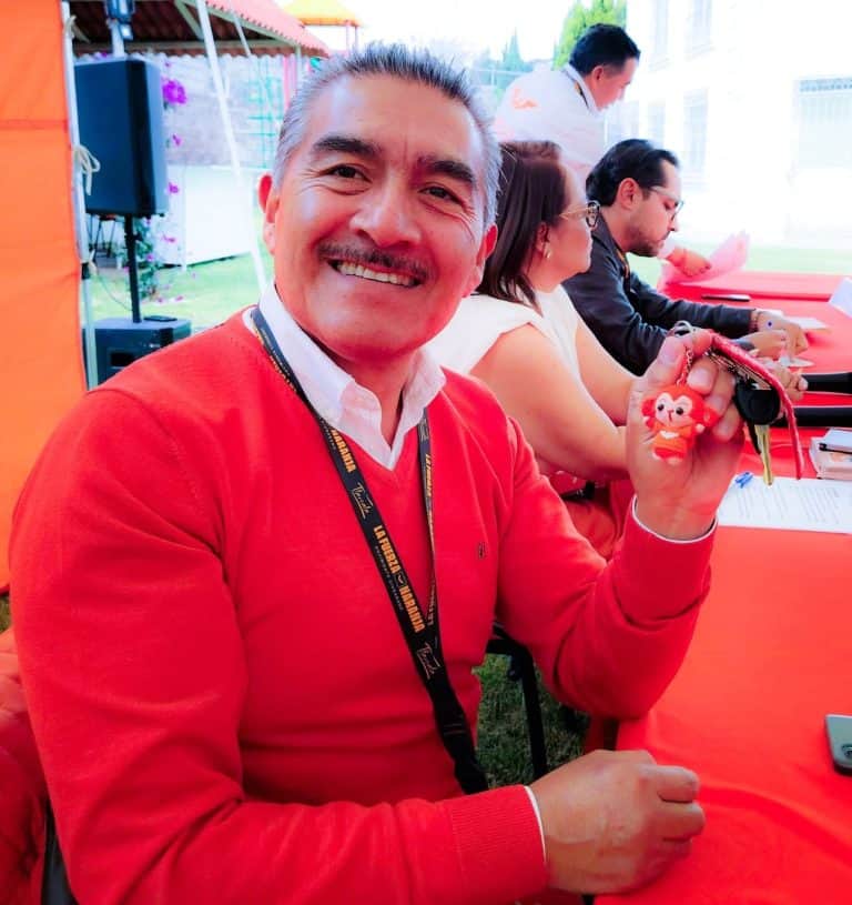 Hombre sonriendo en evento al aire libre, sosteniendo un llavero con figura de oso, con varias personas en el fondo en un ambiente festivo y colorido.