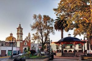 Una plaza en Tlaxcala con árboles, una fuente y un quiosco en el centro, rodeada de edificios históricos y palmeras, durante el atardecer con cielo despejado.