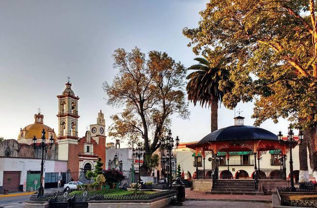 Una plaza en Tlaxcala con árboles, una fuente y un quiosco en el centro, rodeada de edificios históricos y palmeras, durante el atardecer con cielo despejado.