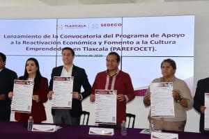 Cuatro personas sosteniendo documentos durante el lanzamiento de la convocatoria del programa de apoyo en Tlaxcala, en un evento oficial con pantalla de fondo.