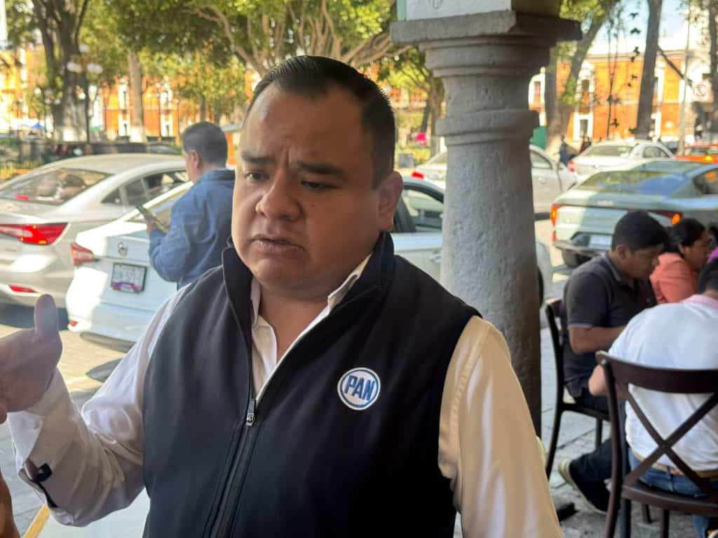 Hombre con chaqueta negra y camisa blanca, con logotipo de PAN, en una calle concurrida con árboles y autos estacionados, parece estar en una conversación o en una reunión al aire libre.