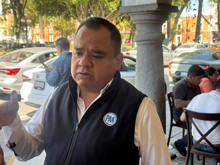 Hombre con chaqueta negra y camisa blanca, con logotipo de PAN, en una calle concurrida con árboles y autos estacionados, parece estar en una conversación o en una reunión al aire libre.