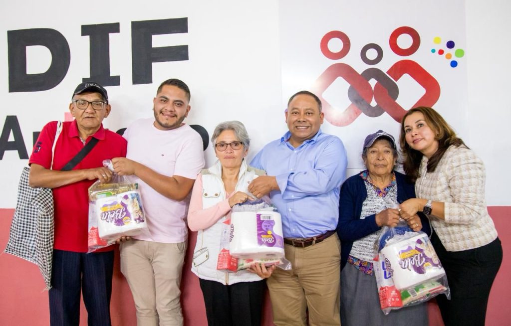 Grupo de seis personas en un evento de entrega de apoyos, sonriendo y sosteniendo paquetes con productos, en un ambiente formal con logotipos y letras en el fondo.