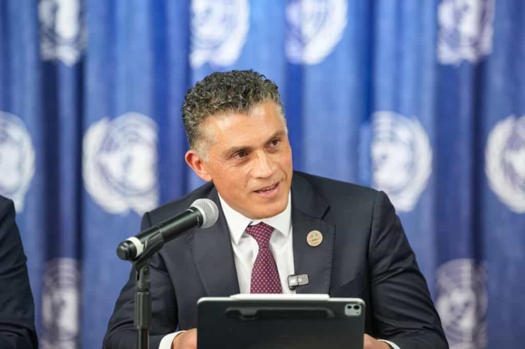 Hombre en traje oscuro hablando en conferencia con fondo de logotipo de la ONU en azul.