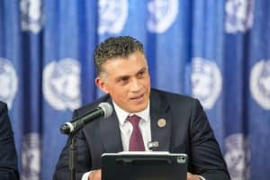 Hombre en traje oscuro hablando en conferencia con fondo de logotipo de la ONU en azul.