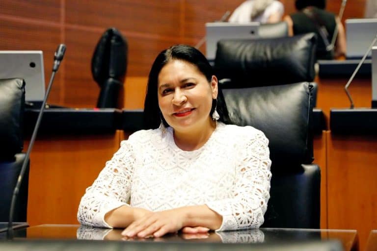 Mujer en una sala de reuniones o conferencia, sentada en una silla de oficina, con fondo de otras personas y mobiliario de oficina.