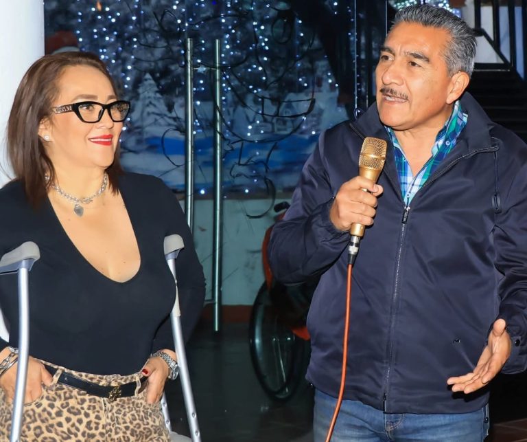 Una mujer con discapacidad en silla de ruedas y un hombre con micrófono en mano conversan en un evento navideño con decoraciones de luces y árbol de Navidad en el fondo.