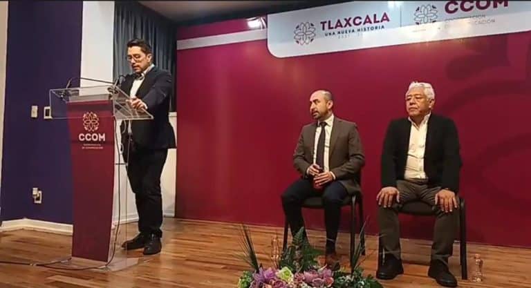 El hombre en traje negro hablando en un podio durante un evento formal en un escenario con fondo rojo y logotipos de Tlaxcala y CCOM.