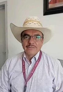 Hombre con gafas y bigote usando sombrero de vaquero y camisa de vestir, sentado en interior con pared blanca y cuadro en el fondo.