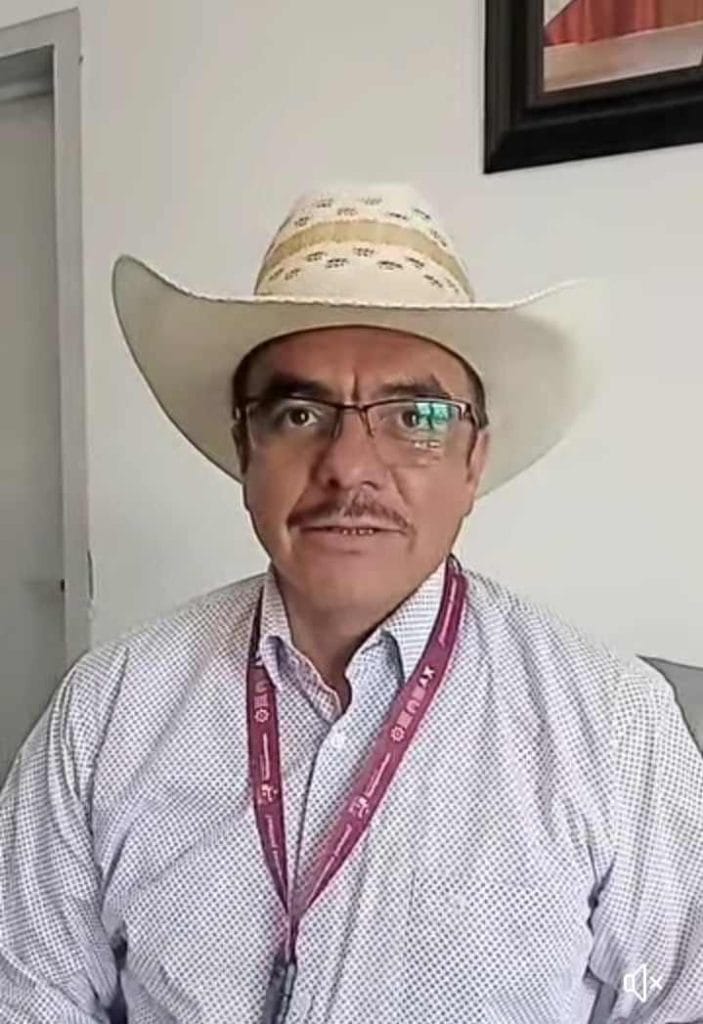 Hombre con gafas y bigote usando sombrero de vaquero y camisa de vestir, sentado en interior con pared blanca y cuadro en el fondo.