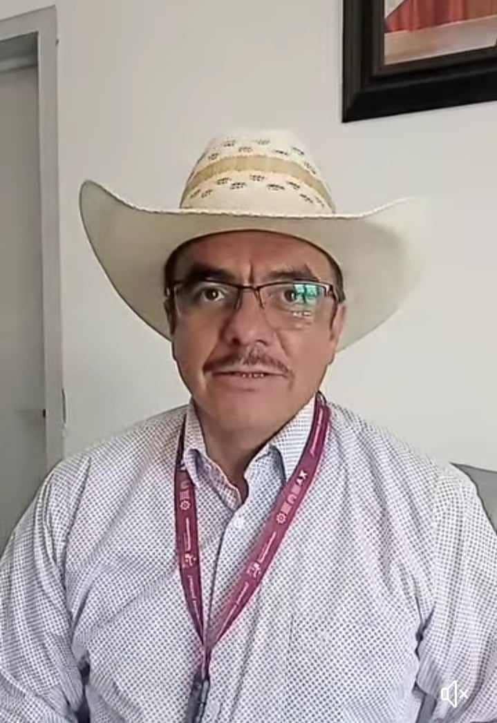 Hombre con gafas y bigote usando sombrero de vaquero y camisa de vestir, sentado en interior con pared blanca y cuadro en el fondo.