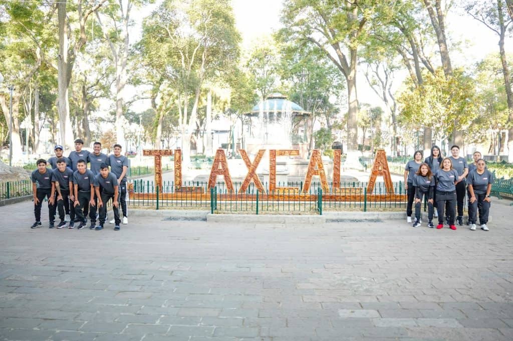 Grupo de jóvenes en Tlaxcala frente al cartel que dice "Tlaxcala" en un parque con árboles y estructura en el fondo.