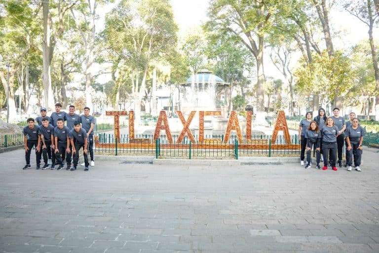 Grupo de jóvenes en Tlaxcala frente al cartel que dice "Tlaxcala" en un parque con árboles y estructura en el fondo.