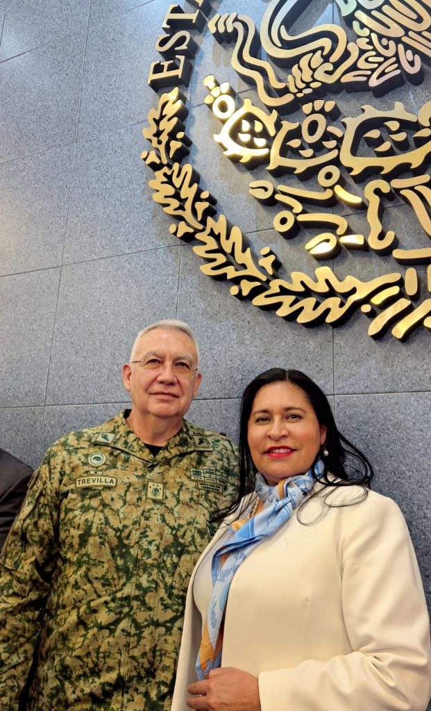 Guerrero en uniforme militar y una mujer con blazer y pañuelo posan juntos frente a un escudo de México en un muro de piedra.