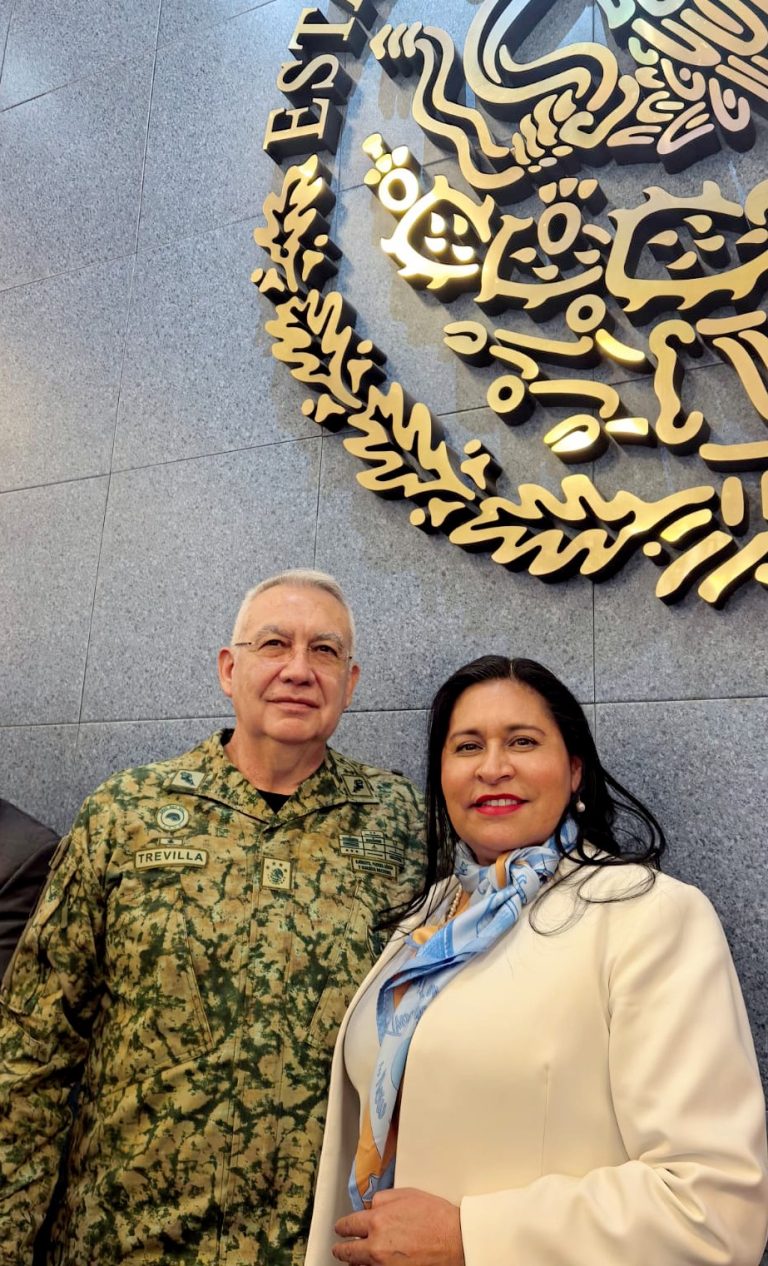 Guerrero en uniforme militar y una mujer con blazer y pañuelo posan juntos frente a un escudo de México en un muro de piedra.
