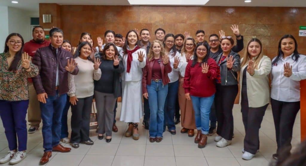 Personas diversas en una reunión, algunas levantando la mano, en un ambiente formal y amigable, en un espacio interior con paredes de color marrón y piso de cerámica.